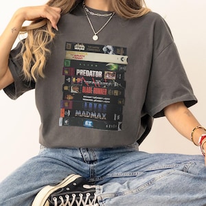 Puede incluir: Camiseta gris oscuro con una pila de cintas VHS vintage con títulos de películas como "Star Wars", "Predator" y "Blade Runner". La camiseta se usa con vaqueros azules y zapatillas negras.