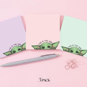Könnte beinhalten: Drei quadratische Haftnotizblöcke in Pastellfarben: Lavendel, Pfirsich und Mintgrün. Jeder Block zeigt ein Cartoon-Bild eines grünen Aliens mit dem Text "THIS IS THE WAY". Ein silberner Stift und rosa Büroklammern sind ebenfalls vorhanden.