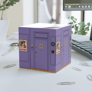 Puede incluir: Un cubo de notas adhesivas morado con un diseño de estilo vintage que presenta puertas, obras de arte enmarcadas y la palabra "MAINA". El cubo está decorado con una estética retro y está sobre una superficie blanca con clips.