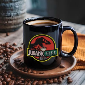 Peut inclure: Une tasse à café en céramique noire avec le logo "Jurassic Perk", représentant une silhouette de dinosaure et une tasse à café. La tasse est remplie de café et posée sur une soucoupe en bois, entourée de grains de café.