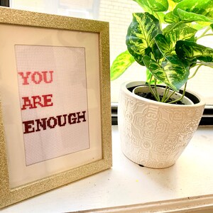 Puede incluir: Un punto de cruz enmarcado con las palabras "YOU ARE ENOUGH" en letras rojas sobre un fondo blanco. El marco es dorado con purpurina.
