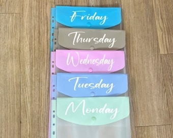 May include: A set of five clear plastic folders with different colored tabs. The tabs are labeled with the days of the week: Monday, Tuesday, Wednesday, Thursday, and Friday. Each tab has a small button.