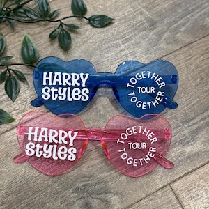 May include: Two pairs of heart-shaped sunglasses. One pair is blue, the other is pink. Both pairs have the text "HARRY STYLES" and "TOGETHER TOUR" in white. The sunglasses are on a wooden surface with green leaves.