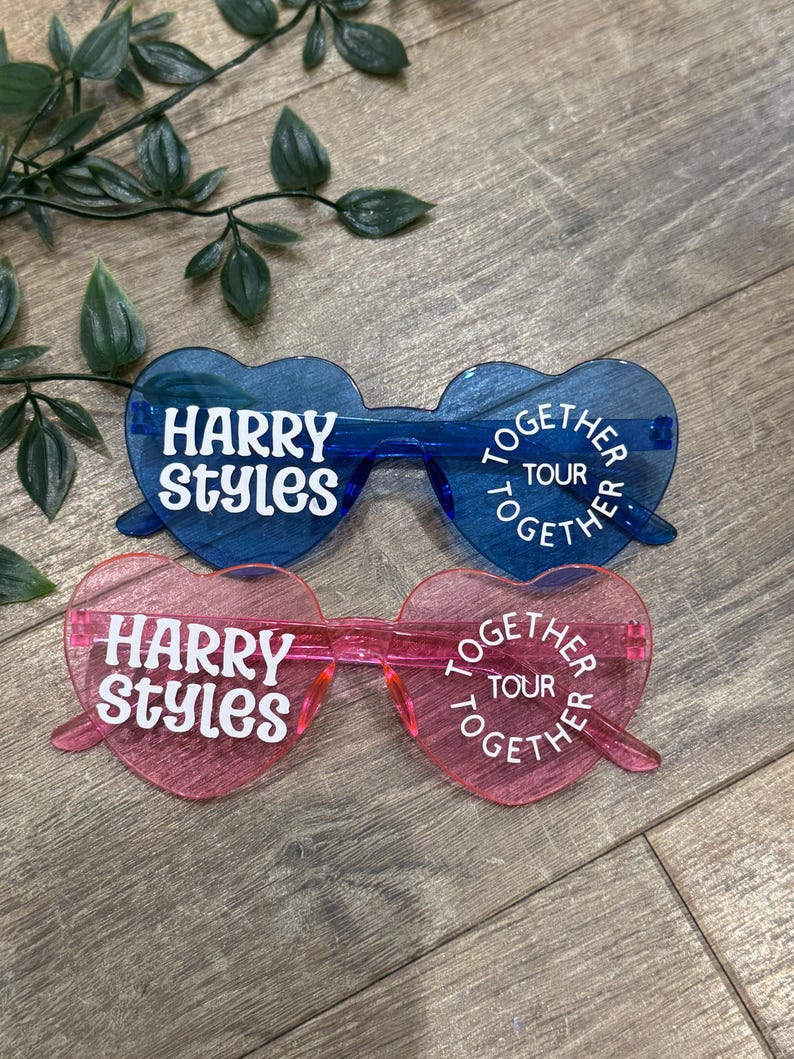 May include: Two pairs of heart-shaped sunglasses. One pair is blue, the other is pink. Both pairs have the text "HARRY STYLES" and "TOGETHER TOUR" in white. The sunglasses are on a wooden surface with green leaves.