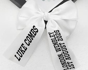 May include: A white satin bow with black text that reads "LUKE COMBS" on one tail and "LONDON WEMBLEY 1ST AUGUST 2026" on the other. The bow is tied in the center and is likely a decorative item.