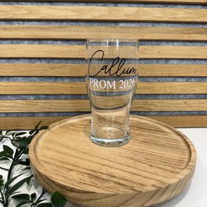 May include: A clear pint glass with the name "Callum" and "PROM 2026" in black script. The glass is on a round wooden tray with a natural wood grain finish. The background has horizontal wooden slats.