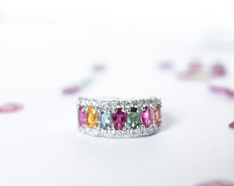 May include: A silver ring with a row of colorful gemstones set in a pave diamond band. The gemstones are in shades of pink, blue, green, and yellow.