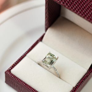 Puede incluir: Un anillo de compromiso de oro blanco con una piedra cuadrada de color verde y pequeños diamantes en la banda. El anillo está en una caja de terciopelo rojo.
