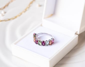 May include: A silver ring with a row of oval-shaped gemstones in shades of pink, green, and orange. The ring is in a white ring box.