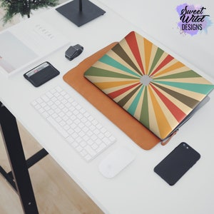May include: A laptop computer with a colorful sunburst design on the cover. The laptop is on a white desk with a keyboard, mouse, and a watch.