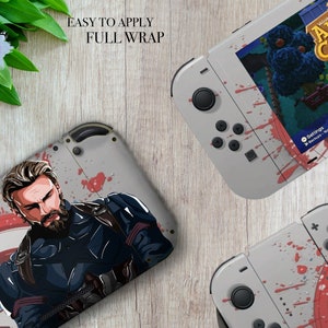 Könnte beinhalten: Eine graue Nintendo Switch Konsolenhaut mit einem rot-weiß-blauen Captain America Design. Die Haut zeigt eine Nahaufnahme von Captain Americas Gesicht und das ikonische Captain America Schild. Die Haut ist einfach anzubringen und bietet eine vollständige Ummantelung für die Konsole.