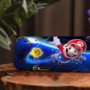 Könnte beinhalten: Eine blaue und schwarze Nintendo Switch-Haut mit einem Cartoon-Bild von Mario, der mit einem gelben Stern durch den Weltraum fliegt. Die Haut hat einen sternenübersäten Hintergrund und den Text "Sweet Wild Designs".