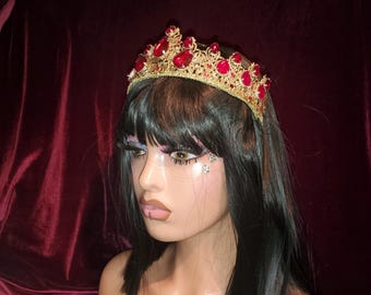 May include: A gold-colored crown with red gemstones. The crown is adorned with multiple red jewels and has an ornate design. The background is a dark red velvet fabric. The crown is a decorative headpiece.
