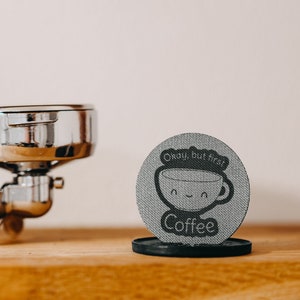 Puede incluir: Un posavasos redondo gris con el texto "Okay, but first coffee" y una taza de café de dibujos animados con una cara sonriente. El posavasos está sobre una superficie de madera junto a un portafiltro de espresso plateado.