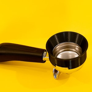 Puede incluir: Un portafiltro de espresso negro y plateado con un accesorio de embudo de silicona negro. El portafiltro está sobre una superficie amarilla.