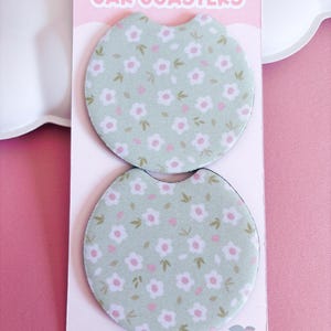 May include: Two car coasters with a floral pattern. The coasters are light green with white and pink flowers and green leaves. The words "CAR COASTERS" are printed at the top of the pink card. The card also says "Designed by @peachylanestudio".