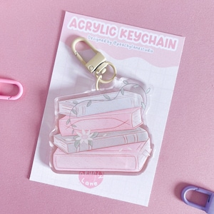May include: Acrylic keychain with a design of a stack of books in pink and grey, decorated with a floral and vine pattern. The keychain has a gold-coloured clasp and is attached to a pink card with the text "Acrylic Keychain".