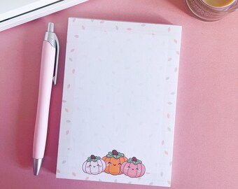May include: A pink pen rests beside a notepad with a pumpkin illustration. The notepad has a border of small leaves and three cartoon pumpkins at the bottom. A white keyboard with pink keys and a candle are in the background.