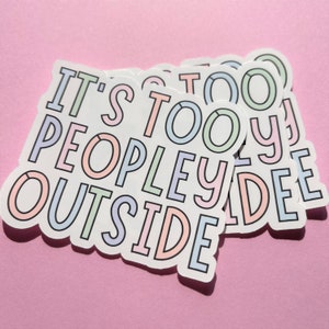 May include: A pile of white stickers featuring the text "IT'S TOO PEOPLE OUTSIDE" in pastel shades with a black border. The stickers are arranged on a pink surface.