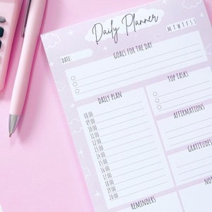 May include: A pink daily planner with the words "Daily Planner" at the top. The planner includes sections for goals, top tasks, affirmations, and gratitude. A pink pen and calculator are in the background.