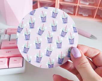 May include: A round coaster featuring a repeating pattern of cartoon bubble tea drinks in purple, blue, and green with red straws. The background is a light pink color with white star-like shapes.