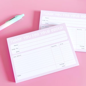 May include: Two light purple "Thank You For Your Order" notepads with white text and lines for order details. A pen with a white and light blue barrel rests on the pink surface. The notepads include fields for name, order number, date, and item details.