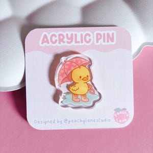 May include: An acrylic pin on a white backing card featuring a yellow duckling wearing pink rain boots and holding a pink polka-dot umbrella while standing in a blue puddle. The card displays the text ACRYLIC PIN and Designed by @peachylanestudio.