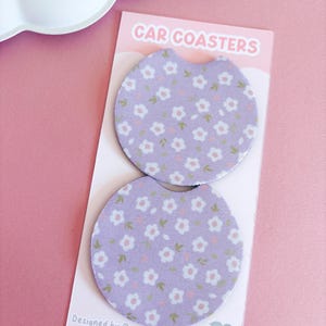 May include: Two car coasters with a floral pattern in a light purple hue. The coasters are round with a curved cutout for easy removal. The packaging reads "CAR COASTERS" and "Designed by @peachylanestudio."