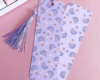 May include: Two lavender bookmarks with a repeating pattern of potion bottles, stars, and crescent moons. One bookmark has a lavender tassel. The bookmarks are on a pink surface, near a white keyboard.