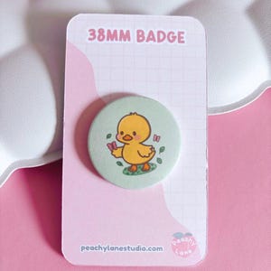 May include: A circular 38mm badge featuring a cartoon yellow duckling on a light green background. The duckling holds a small pink butterfly. The badge is mounted on a rectangular white and pink card with the text 38MM BADGE and peachylanestudio.com.