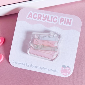 May include: An acrylic pin featuring a stack of books in shades of pink, gray, and blue, adorned with delicate floral and vine details. The pin is on a pink and white card that reads "ACRYLIC PIN" and "Designed by @peachylanestudio."