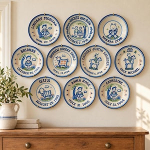 May include: A collection of nine round ceramic decorative plates arranged on a wall, each featuring blue hand-painted illustrations of children, animals, and names with birth dates including Lindsay Morgan, Jackie and Ron, and Hannah Lynn.