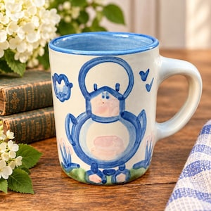 May include: A ceramic mug with a white base and a hand-painted blue duck illustration. The design features a stylized blue duck with pink accents on the chest and feet, surrounded by small blue clouds and birds. The rim is painted in blue.