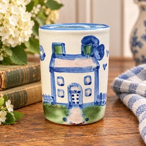 May include: A cylindrical ceramic tumbler featuring a hand-painted blue and white house illustration. The design includes a central house with a blue roof, windows, and door, set against a green ground with small blue hearts and cloud accents.