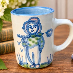 Op de afbeelding: Een keramische mok met een witte basis en een blauwe rand, voorzien van een handgeschilderde blauwe en groene illustratie van een persoon met een hoed en sjaal die een fluit vasthoudt. Het ontwerp bevat kleine blauwe vogels en bloemmotieven.