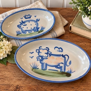 May include: Two oval ceramic platters with a cream-colored base and a blue rim. One platter features a blue hand-painted pig design, while the other displays a blue and light pink hand-painted cow design, both standing on green patches.