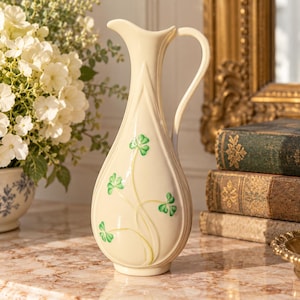 May include: A cream-colored ceramic pitcher vase with a slender neck, a curved handle, and a pointed spout. The body features embossed vertical lines and a painted green shamrock design with delicate stems extending upward from the base.