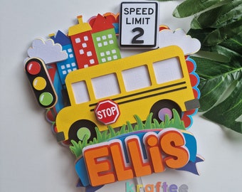 May include: A yellow school bus cake topper with a stop sign, a traffic light, and a speed limit sign that says "Speed Limit 2". The topper also includes a city skyline and the name "Ellis" in orange letters.
