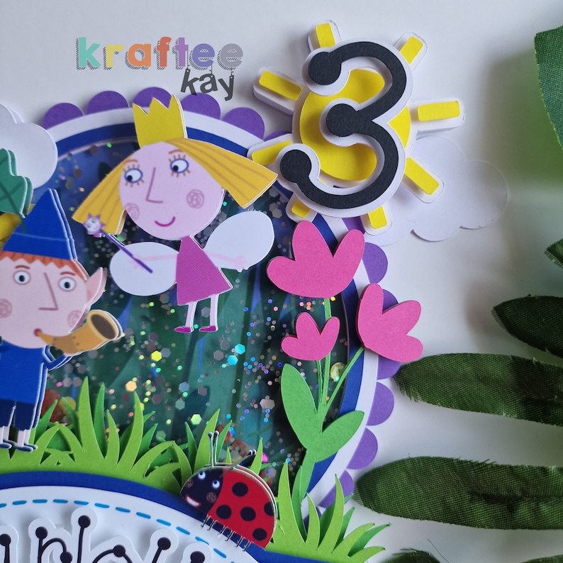 May include: A 3D birthday cake topper featuring Ben & Holly characters. The topper includes a yellow sun with the number 3, a ladybug, a flower, and a green background with glitter.