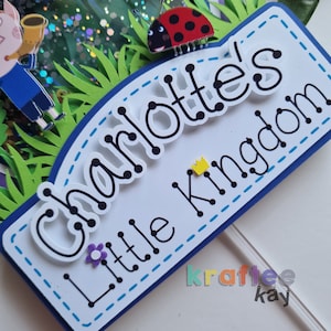 May include: A white cake topper with the text "Charlotte's Little Kingdom" in black lettering with a yellow crown and a ladybug. The topper is decorated with blue and white stitching.