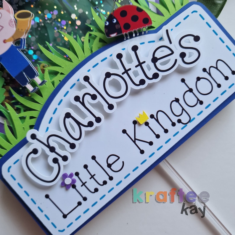 May include: A white cake topper with the text "Charlotte's Little Kingdom" in black lettering with a yellow crown and a ladybug. The topper is decorated with blue and white stitching.