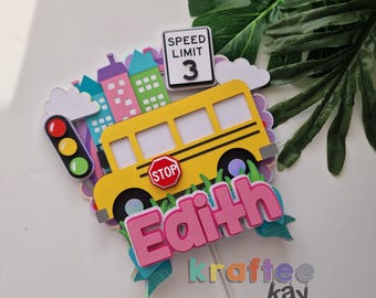 May include: A colorful cake topper featuring a yellow school bus, a stop sign, and a speed limit sign. The name "Edith" is displayed in pink letters. The topper includes a cityscape, clouds, and a traffic light.