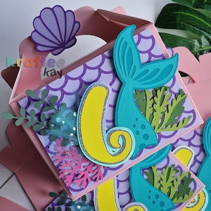 May include: A pink and purple mermaid-themed party favor box with a teal tail, yellow number six, and green seaweed decorations.