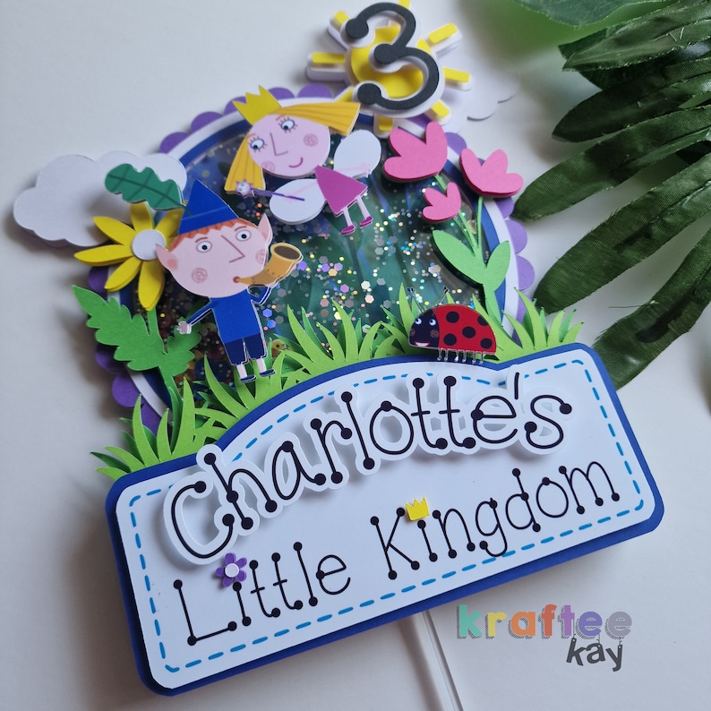 May include: A cake topper featuring Ben & Holly's Little Kingdom characters, Ben and Holly, with a glittery background and a sign that reads "Charlotte's Little Kingdom".