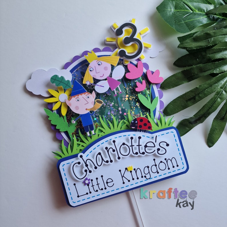 May include: A colorful cake topper featuring characters from the children's show 'Ben & Holly's Little Kingdom'. The topper includes Ben, Holly, a ladybug, flowers, and a sun with the number 3. The topper also includes a sign that says 'Charlotte's Little Kingdom'.