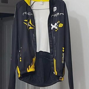 Puede incluir: Chaqueta de carreras negra y amarilla con detalles blancos. La chaqueta tiene cierre de cremallera y presenta varios logotipos y texto, incluyendo "ROCKRIDER" en el cuello. El diseño incorpora formas geométricas y la palabra "RACING".