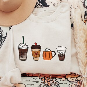 May include: A white sweatshirt with a graphic of four different coffee drinks. The drinks are a caramel macchiato, a pumpkin spice latte, a pumpkin cream cold brew, and an iced coffee. The text "Taryn Lora Designs" is at the bottom of the graphic.