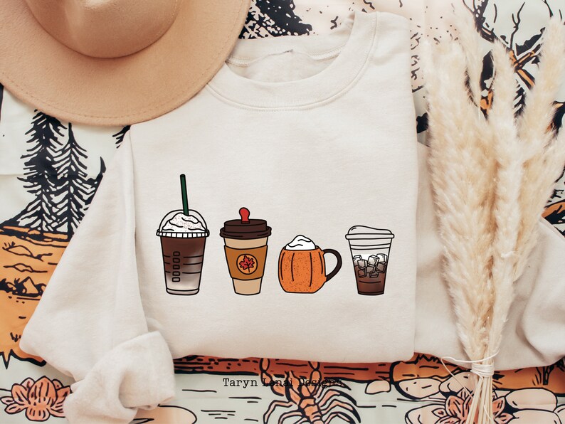 May include: A white sweatshirt with a graphic of four different coffee drinks. The drinks are a caramel macchiato, a pumpkin spice latte, a pumpkin cream cold brew, and an iced coffee. The text "Taryn Lora Designs" is at the bottom of the graphic.
