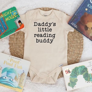 May include: A beige baby onesie with the text "Daddy's little reading buddy" printed in black. The onesie is surrounded by five children's books, including "Goodnight Moon", "The Very Hungry Caterpillar", "Pout-Pout Fish", and "Snuggle Tight, Kiss Good Night".