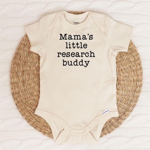 May include: A cream-colored baby onesie with short sleeves and the text "Mama's little research buddy" printed in black.